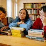 Three diverse students studying together in a university library with books around them.